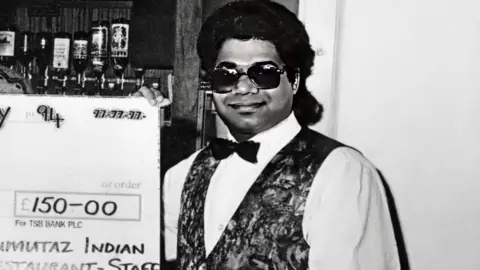 Ken Amer/Firecrest Films Black and white photograph of a man in a restaurant holding a big cheque. He is smiling. The man has long, dark, swept back hair. He is wearing a white shirt, black bow tie and a patterned waistcoat.