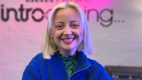 Caswell, inside the BBC Music Introducing studio, wearing a bright blue fleece zip jacket and vibrant red lipstick