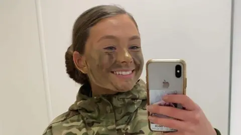 Family Handout A mirror selfie of Gunner Beck wearing her camouflage military uniform, with her hair tied back into a slick low bun. She has camouflage green and brown face paint on and is smiling at the mirror.