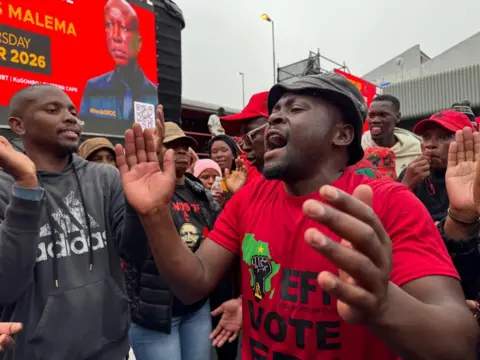 A man in a red EFF T-shirt is among a crowd of Malema supporters. He can be seen chanting and holding his hands out to clap.