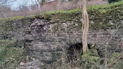 A brick retaining wall on the A684 at Smorthwaite which has partially collapsed. The wall also includes a culvert, with an arched structure. There is moss on part of the wall and bricks on the ground.