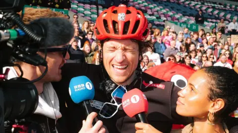 A man in a red helmet and cycling gear is in tears. There is a man one side of him holding a blue microphone and a woman the other side of him holding a red microphone. Behind him, a crowd of people sit in a open air stadium.