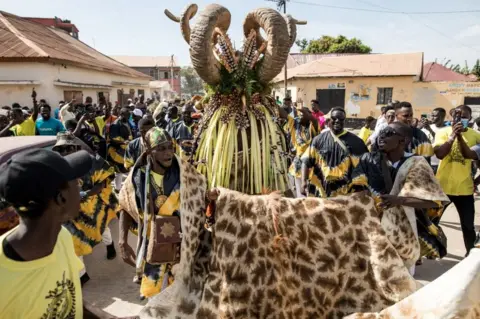 AFP Members of the 'Ekum Baba' escort their hunting costume during the annual hunting festival in Banjul on January 2, 2023. - This hunting festival occurs every new years in Banjul. The two groups 'Ekum Baba' and 'Odilleh' come to the streets of Banjul every year to battle for the title of best animal heads,masques and costumes