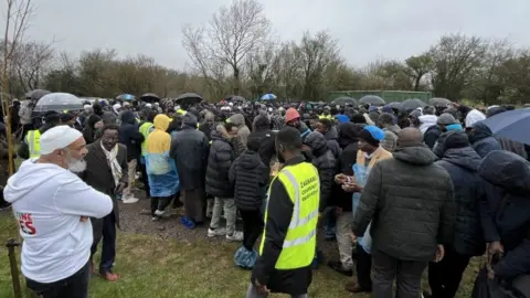 A large group of people gathered to watch a funeral, many carrying umbrellas