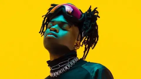 Jordan Adetunji, a black man in his 20s, poses wit his eyes closed against a bright yellow backdrop. Jordan, who has his braided hair tied up, wears ski goggles on top of his head, a high necked black top and two chunky silver chains around his neck. He looks up, slightly angled to the left of the camera, with his face lit by a blue/ green light