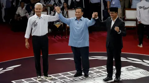 Reuters Ganjar Pranowo, Prabowo Subianto, Anies Baswedan hold hands as they attend a televised debate at the election commission headquarters in Jakarta, Indonesia, December 12, 2023.