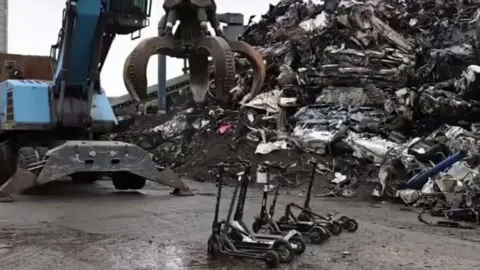 Northamptonshire Police E-scooters lined up to be grabbed by a blue mechanical claw next to a pile of scrap metal