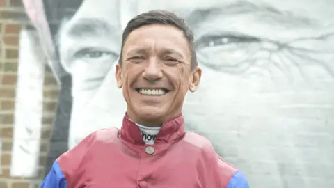 Jamie Niblock/BBC Frankie Dettori smiling in front of a monochrome mural of himself wearing a horseriding helmet