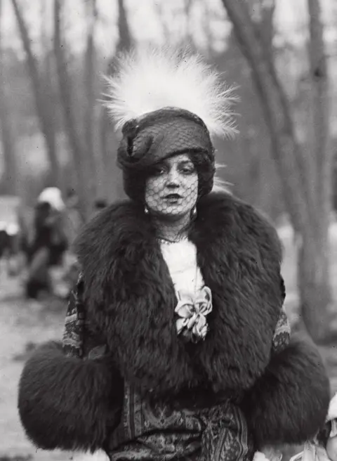  Jacques Henri Lartigue A portrait of a woman in a park wearing an elaborate hat, face veil and thick fur coat