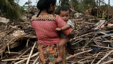 AFP via Getty Images A Rohingya woman carries her baby next to her destroyed house at refugee camp in Sittwe, Myanmar. Photo: 16 May 2023