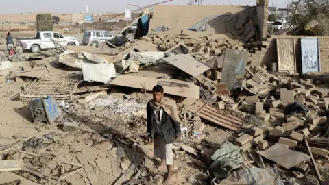 Reuters A man stands at the site of a reported Saudi-led coalition air strike on the outskirts of the rebel-controlled northern Yemeni city of Saada (22 January 2018)