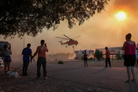 Nicolas Economou / Reuters People look at a firefighting helicopter filling water from a pool, as a wildfire burns in the village of Gennadi, on Rhodes