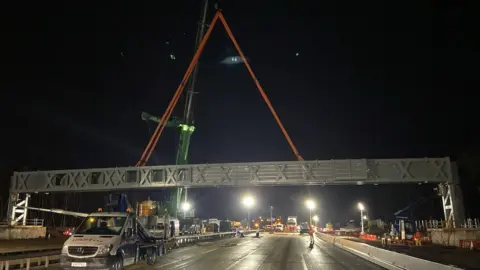 National Highways: South-East A night time image shows a gantry being lifted into place above the M25