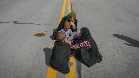 Afghan people sit along the tarmac as they wait to leave the Kabul airport