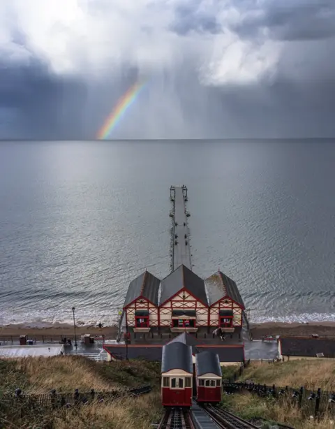 Nicky Hunter View down a vertical railway to a pier jutting out in to the sea and rainbow in the distance