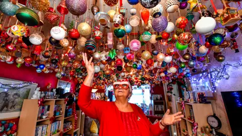 PA Media Nanna Baubles standing underneath hundreds of Christmas baubles hanging from her living room ceiling