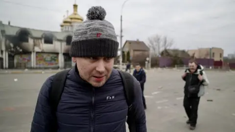 Orla Guerin/BBC Andrei is a man in his 30s, pictured wearing a backpack and a bobble hat as he left the town of Irpin