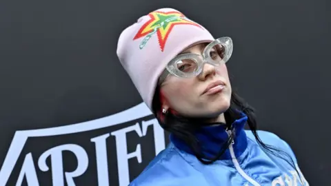 Getty Images Billie Eilish attends the 2023 Variety Hitmakers Brunch at NYA WEST on December 02, 2023 in Hollywood. Billie, a 21-year-old woman, wears a pink beanie hat with a green, yellow and red star on the front. She wears thick rimmed clear glasses and a blue zip-front jacket. She's pictured in front of a black background with 'Variety' printed on it.