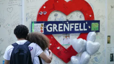 PA Media Two people hug in front of a sign for Grenfell