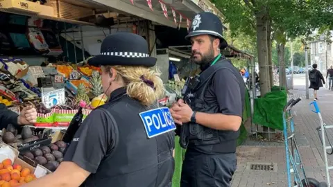 Cambridgeshire Constabulary Two police officers, one male and the other female. They are standing next to a fruit stall. The officers are facing towards the stall.