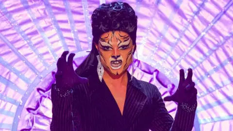 BBC/World of Wonder/Guy Levy Gothy Kendoll on the Ru Paul's Drag Race UK runway in 2019. Gothy wears a black pinstripe suit and black wig and has painted her face to resemble a tiger. She holds her hands up gesticulating a roar.