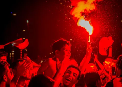 NurPhoto/Getty Images Algeria fans smile and gesture in Bordeaux's Place de la Victoire in Bordeaux. One person holds a flare up, turning the air red.