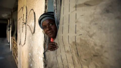 BBC/SHIRAAZ MOHAMED An unidentified woman peeks out of her room. With the doors and door frames removed, a fabric sheet serves as a makeshift door in the derelict San Jose building in Johannesburg, South Africa