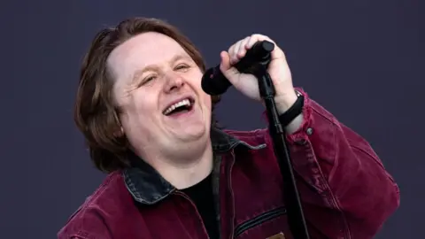 Getty Images Lewis Capaldi on stage during Radio 1's Big Weekend in Dundee in 2023. Lewis is a 27-year-old white man with brown hair almost to his shoulders. He wears a burgundy jacket with a black collar and smiles as he sings into a microphone, which he holds toward him with his left hand. He is pictured on stage in front of a dark grey backdrop