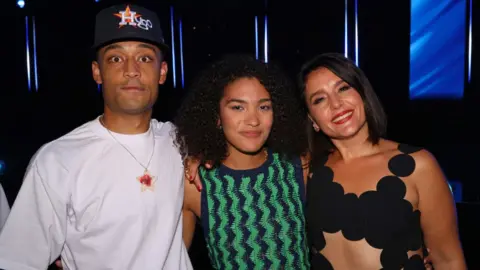 Getty Images Loyle Carner, Olivia Dean and Jessie Ware attend The Mercury Prize 2023 awards show at Eventim Apollo on September 07, 2023 in London. Loyle wears a white T-shirt and black cap emblazoned with 'Hugo' and looks wide-eyed at the camera. Olivia, about a foot shorter, stands next to him, she wears her dark curl hair loose and smiles at the camera. She wears a green and blue zig-zag patterned dress. To her right is Jessie Ware, who tilts her head towards Olivia. She has short, straight dark hair and smiles at the camera, wearing deep red lipstick. Her dress is formed of connecting black spots. The trio are pictured inside at the ceremony and it is dark behind them with the exception of a blue screen