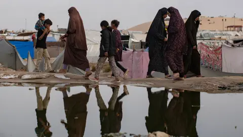 People are reflected as they walk in front of a sewage spill in a makeshift camp on the outskirts of Rafah on 26 April