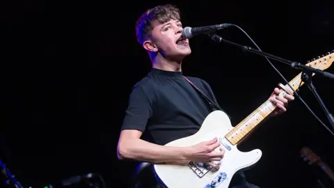 Getty Images Musician Tom A Smith playing at a gig. He holds a white guitar and looks emotional, his mouth open wide as as he sings into a microphone. He's wearing a dark t-shirt and glancing out of the side of his eye at the camera.