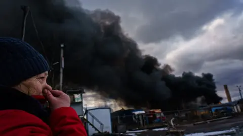 Getty Images A woman holds her nose to prevent inhaling the smell of chemicals caused by the plume of smoke from the fire caused by a Russian bombardment of a storage facility on the edge of Kalynivka, 8 March 2022.