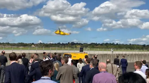 A yellow helicopter in the sky with a small crowd watching