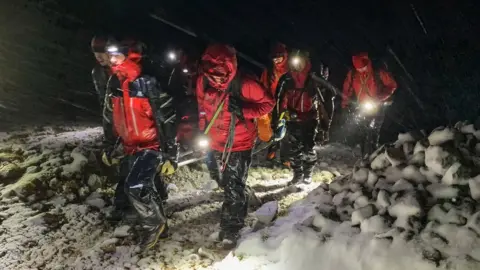 Keswick Mountain Rescue A group of volunteers in red and black waterproofs walk at night, lit by head torches, in sleet and snow