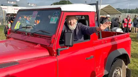 Michael Eavis in a red Land Rover at the entrance to Glastonbury Festival