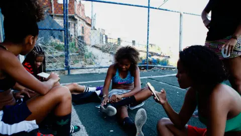 Gustavo Oliveira/WBR Photo One of the girls, centre, fixes her shoe as the girls talk and check their phones after training