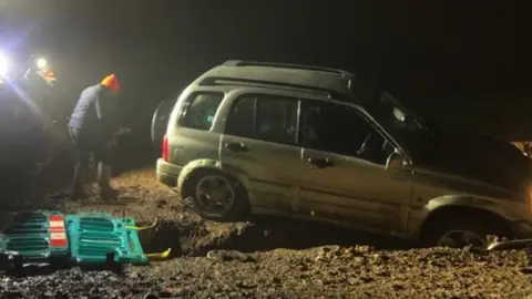 Jon King Photo of a large silver car abandoned in a trench. The front driver's side wheel is in the ditch. Two people can be seen behind. It is dark - captured at night.