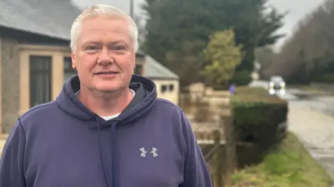 Martin is standing outside in front of a house. There is a visible flood to the right of the image and a car attempting to drive through it. He has white hair and is wearing a blue jumper with a hood.