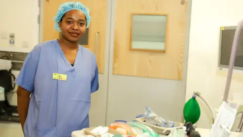 The image shows a black woman wearing scrubs and a hair net. She is in a medical room and different medical instruments can be seen on a table in front of her