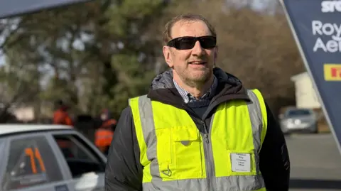 A man wearing a yellow hi-vis jacket and sunglasses stands in a car park.
