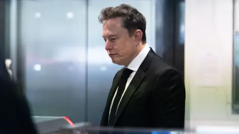 Musk, wearing a black suit, white shirt and black tie, looks down toward the floor while waiting to pass through security at a courthouse in California.