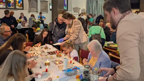 Children and adults sit around a white table crafting and painting eggs with some small candles. They appear to be in a church or church hall