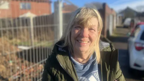 Photograph of 67 year-old Beverley Simms from Oldham. She is pictured in front of a building site in Derker.