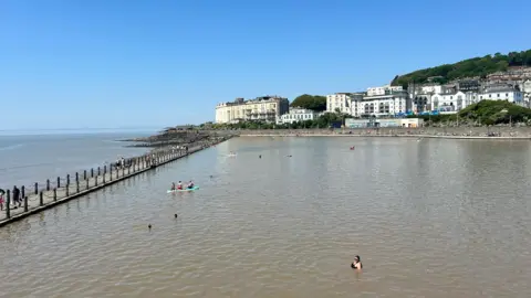 A large marine lake with people swimming and paddle boarding in it. The water is a murky brown colour, and the sky is blue, with old hotels and houses lining the promenade in the distance. 