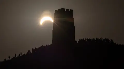 The church tower at the top of Glastonbury Tor with lots of people surrounding it in darkness with the partial solar eclipse visible in the sky
