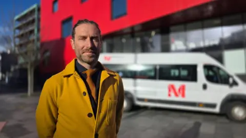 A man in a mustard coloured coat and orange tie and black t-shirt. He is standing in front of a large red building and a school bus. 