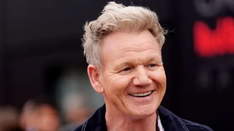 Gordon Ramsay looking to the side of the camera and smiling. The background behind him is blurred.