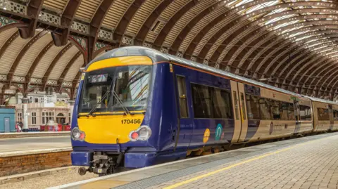  A train stands at a railway station platform covered by a historic curving iron canopy. Architectural columns line the platforms and a station pub is on the far platform.