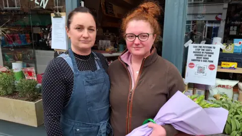 BBC/Seb Cheer Ruth Phillips and Charlotte Kneafsey stand outside a shop looking at the camera. Ruth is wearing a polka-dot T-shirt under a denim apron, while Charlotte is wearing a pink shirt under a brown fleece. Charlotte is also carrying a bouquet of flowers.