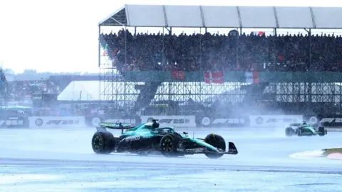 Reuters A Formula 1 racing car in the rain at Silverstone. Behind the car is a grandstand full of fans.